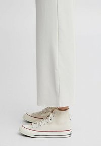 Sneakers alte in tela color crema con suola in gomma, lacci bianchi e dettagli rossi e blu scuro. Abbinati a pantaloni bianchi larghi.