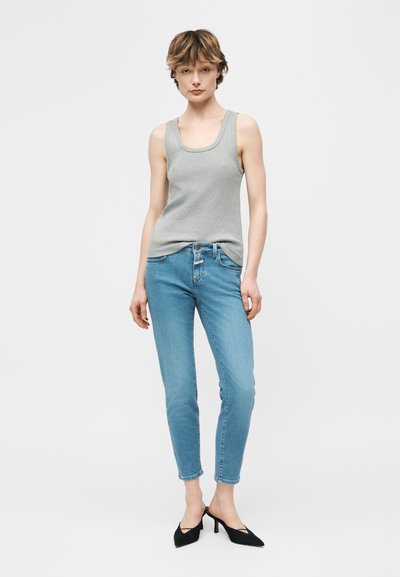 Femme portant un haut gris sans manches, un jean slim bleu clair et des talons noirs à bout pointu, debout devant un fond blanc uni.