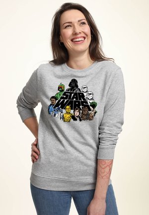 Kobieta uśmiechająca się, ubrana w szarą bluzę z postaciami i logo Star Wars, stoi z jedną ręką na biodrze na jednolitym tle.