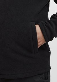 Pullover in pile nero con una superficie testurizzata, dotato di una tasca laterale e polsini aderenti. Senza loghi o motivi visibili.