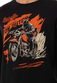 T-shirt en coton noir avec un graphisme d'un motard en orange et blanc, accompagné du texte "California Von Dutch" et d'éléments de design dynamiques.