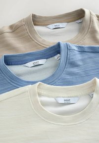 Trois t-shirts à col rond de coupe décontractée en beige, bleu clair et crème, présentant une texture côtelée et des étiquettes de marque visibles au niveau du col.