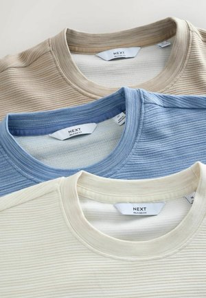 Trois t-shirts à col rond de coupe décontractée en beige, bleu clair et crème, présentant une texture côtelée et des étiquettes de marque visibles au niveau du col.