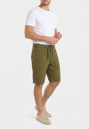 Uomo che indossa una t-shirt bianca a maniche corte, pantaloncini verde oliva con coulisse e tasche, e scarpe da ginnastica beige, in piedi davanti a uno sfondo semplice.