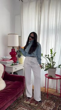 Aurinkolasit päässään oleva nainen, jossa on lyhyt farkkutakki ja valkoiset housut, seisoo valoisassa huoneessa, jossa on punainen sohva, valkoiset verhot ja viherkasvi punaisella pöydällä.