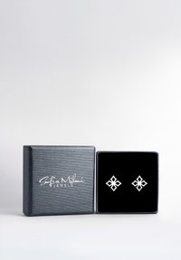 Silvriga geometriska örh ängen visade i en smyckesask med svart sammet. Asken har en strukturerad mörkgrå yta och en vit logotyp.