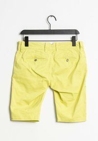 Shorts en coton jaune avec deux poches arrière, boutons en métal et un design décontracté. Suspendus à un cintre en plastique noir sur fond blanc.