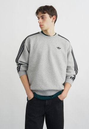 Junger Mann mit Händen in den Taschen, der einen grauen Adidas-Sweatshirt mit schwarzen Streifen an den Ärmeln und schwarzen Hosen trägt, schaut zur Seite.