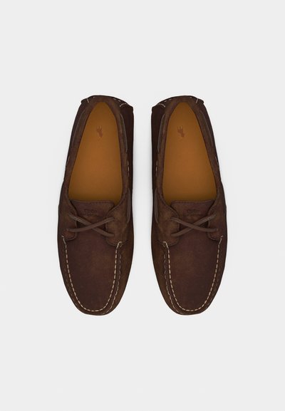 Ζευγάρι καφέ σουέντ μοκασίνια με δερμάτινα κορδόνια και αντίθετη λευκή ραφή, απεικονισμένα από πάνω σε λευκό φόντο.