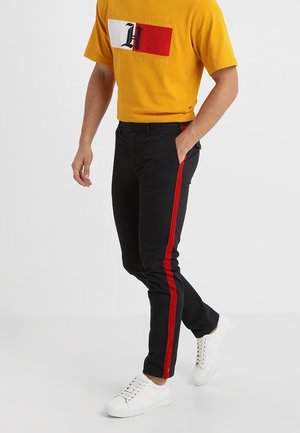Pantaloni neri slim-fit con strisce rosse laterali, realizzati in un mix di cotone, abbinati a una t-shirt gialla e sneaker bianche. Texture liscia.