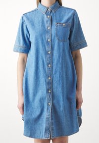 Abito camicia in denim, azzurro chiaro, maniche corte, chiusura frontale con bottoni, taschino sul petto e orlo curvo. Il tessuto ha una consistenza liscia e morbida.
