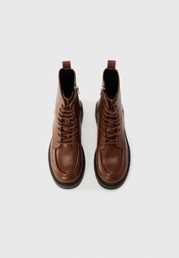 Bottines en cuir marron avec une texture lisse, à lacets à l'avant, zips latéraux et une épaisse semelle noire. Bout plat et forme arrondie.