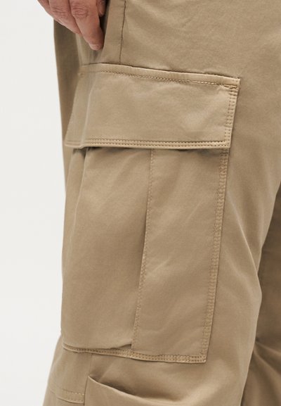 Κοντινό πλάνο ενός χεριού κοντά σε μια μεγάλη μπεζ τσέπη φορτίου σε μπεζ παντελόνι, που δείχνει λεπτομερή ράψιμο και υφή υφάσματος.