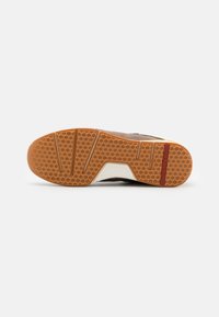 Semelle de chaussure en nubuck brun avec un motif en nid d'abeille texturé, présentant un accent blanc et une bande rouge, conçue pour l'adhérence et la stabilité.
