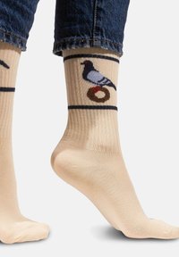 Beżowe skarpetki o prążkowanej fakturze, z rysunkowym gołębiem siedzącym na brązowym okręgu, otoczonym ciemnym paskiem u góry.