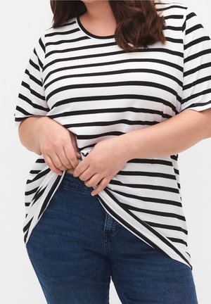 Femme portant un haut à rayures noires et blanches à manches courtes et un jean bleu, ajustant l'ourlet de son haut avec les deux mains devant un fond blanc.