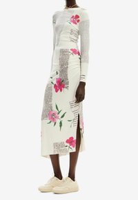 Robe à manches longues blanche avec motifs floraux et impression de texte. Présente une fente sur le côté et des détails froncés. Associée à des baskets de couleur claire.