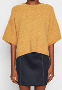 Senapsgul oversized stickad tröja med korta ärmar och en lös passform, kombinerad med en svart minikjol med synliga sömnadsdetaljer.