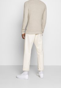 Man wearing a beige long-sleeve sweater, off-white rolled-up pants, white socks, and white sneakers, standing facing away.