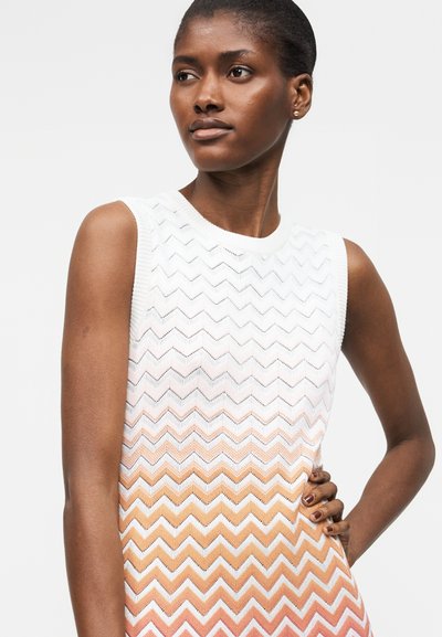 Γυναίκα με κοντά μαλλιά που φορά ασπρό αμάνικο τοπ με πορτοκαλί ζιγκ-ζαγκ σχέδιο, ποζάρει με το ένα χέρι στη μέση.