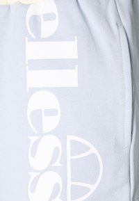 Tissu bleu clair avec grand texte de la marque « ellesse » en blanc et logo, ainsi qu'une poche latérale visible sur le côté droit.