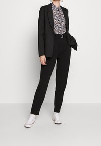 Blazer nero sopra una blusa a fiori, abbinato a pantaloni neri a vita alta e sneaker bianche. Design semplice e sartoriale.