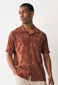 Chemise à manches courtes en tissu texturé terracotta, dotée d'un motif orné de soleils et de feuilles. Comprend une poche poitrine et une fermeture à boutons.