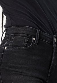 Sorte højtaljede jeans med en tætsiddende pasform, der har forlommer og diskrete syninger, præsenteret sammen med en sort top.
