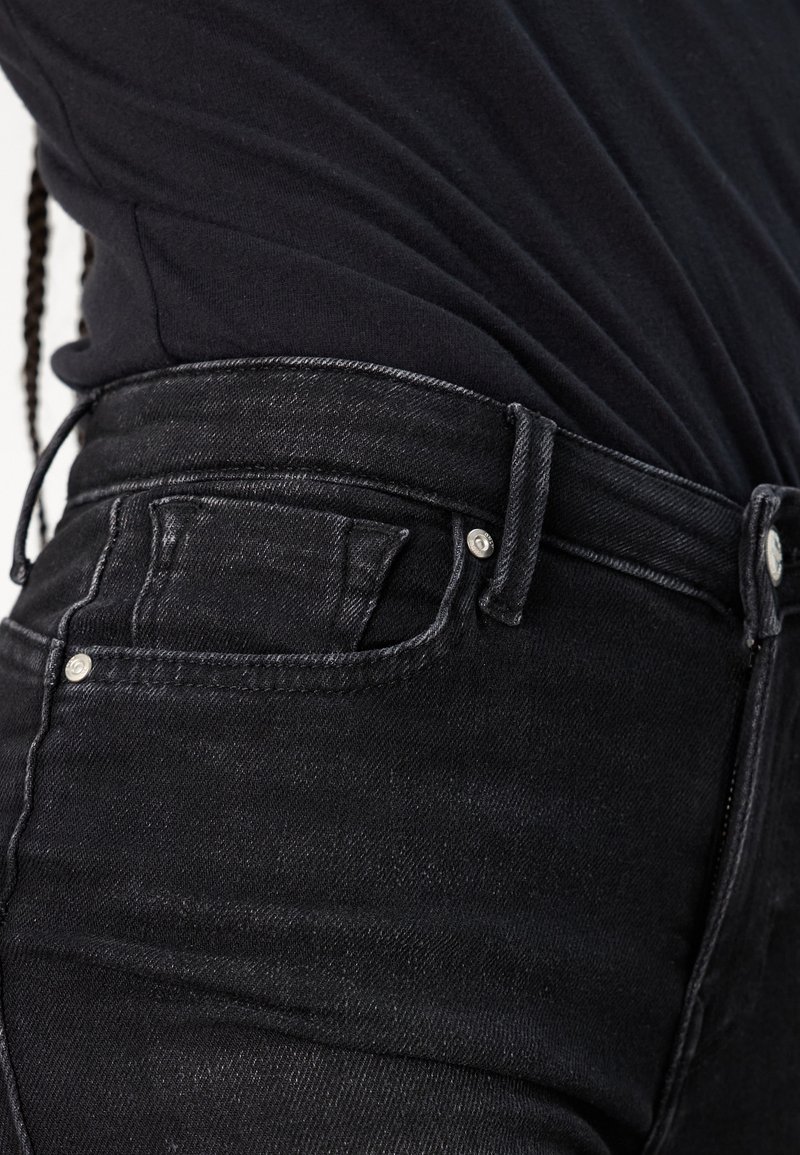 Jean noir taille haute avec un design ajusté, featuring des poches avant et des détails de coutures subtils, présentés aux côtés d'un haut noir.