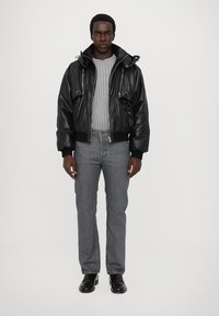 Veste bombardier en cuir noir avec une fermeture éclair à l'avant, des poignets côtelés et une capuche, associée à un pull à côtes gris et un jean gris. Chaussures noires.
