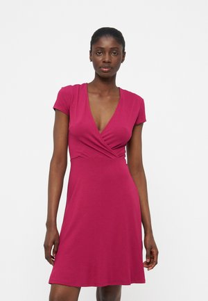 Γυναίκα όρθια, φορώντας ένα μαγνητικό φόρεμα με κοντά μανίκια, βαθύ ντεκολτέ σε σχήμα V και μήκος μέχρι το γόνατο, σε απλό λευκό φόντο.