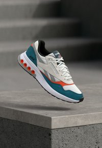 Białe, turkusowe i pomarańczowe sneakersy Reebok z designem na sznurówki unoszące się nad betonowym krawędzią, z rozmytymi schodami w tle.