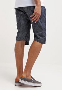 Pantalones cortos de color azul marino con un ajuste relajado, con un diseño geométrico, un corte a la rodilla y un tejido ligero. Llevados con zapatos grises tipo slip-on.