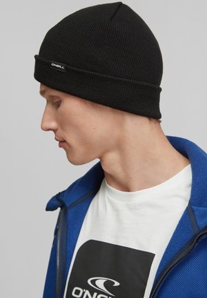 Black ribbed beanie with a folded cuff, featuring a small white logo tag. The model wears a blue zipped jacket and a white t-shirt.