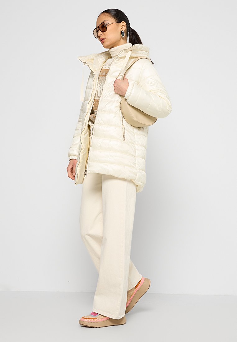 Giacca cremosa imbottita con cappuccio, finitura testurizzata e dettagli con zipper, abbinata a pantaloni a gamba larga color crema e scarpe da ginnastica colorate.