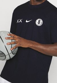 Camiseta de algodón azul marino con un logo blanco de Nike y un gráfico de los Brooklyn Nets. Un jugador sostiene un balón de baloncesto gris con acentos negros.