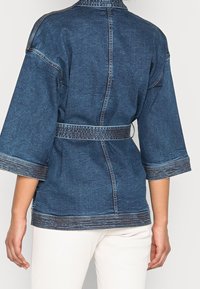 Kurtka dżinsowa wyróżniająca się głębokim niebieskim kolorem, szerokimi rękawami i paskiem w talii. Posiada kontrastujące detale w szwach oraz teksturowany materiał.