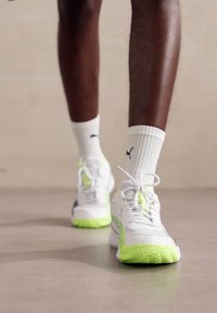 Zapatillas deportivas blancas con acentos verdes y suelas texturizadas, que cuentan con cordones y un diseño elegante. Llevadas con calcetines blancos hasta el tobillo.