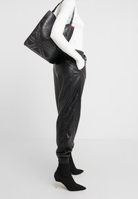 Czarne skórzane spodnie z bocznymi szwami, biała, prążkowana bluzka z długim rękawem oraz czarna torba na ramię z ćwiekowym wzorem i czerwonymi akcentami.