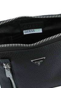 Bolsa negra con cremallera hecha de cuero texturizado, con una cremallera en tono plateado y una etiqueta de logo. El interior incluye una etiqueta GUESS ECO.