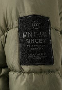 Veste doudoune vert olive avec une étiquette en tissu noir texturé comportant du texte en relief : "MNT SINCE 97" et des instructions d'entretien.