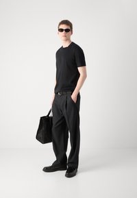 T-shirt nera a maniche corte, pantaloni neri sartoriali, scarpe nere lucide, cintura nera, borsa nera strutturata e occhiali da sole neri sottili.