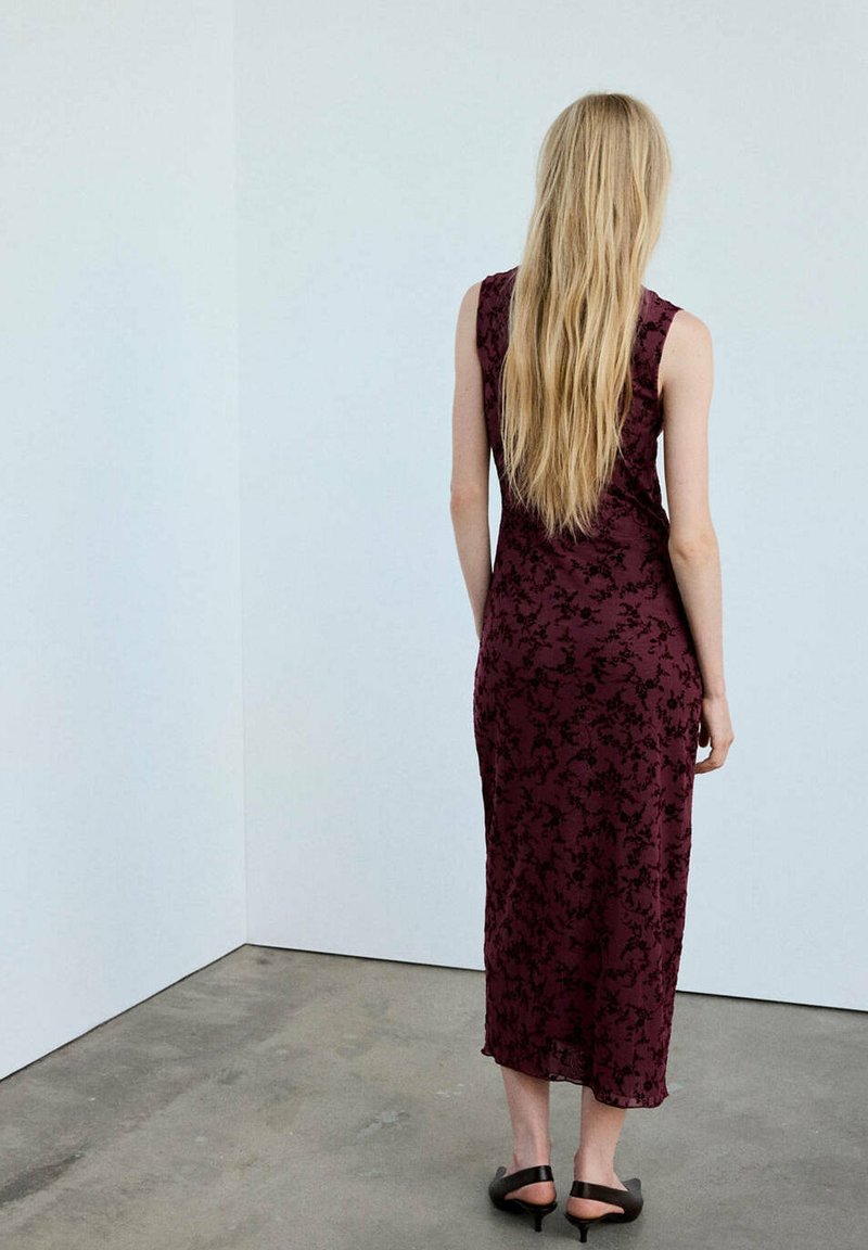 Femme aux longs cheveux blonds portant une robe fleurie rouge foncé sans manches et des sandales noires, debout face à un mur d'angle blanc.