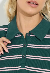 Zielona polo z kołnierzykiem, o ryflowanej fakturze, z różowymi poziomymi paskami i dwoma guzikami przy szyi.