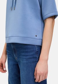 Jasnoniebieska bluzka z krótkim rękawem, okrągłym dekoltem i bocznym rozcięciem, w zestawie z ciemnoniebieskimi dżinsami. Wyraźny metalowy logo akcent w pobliżu dolnego brzegu.