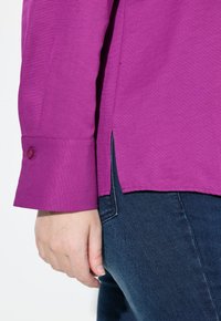 Camicia lunga viola con polsini abbottonati, fessure laterali all'orlo, mostrata con jeans in denim scuro. Il tessuto appare liscio e ben strutturato.