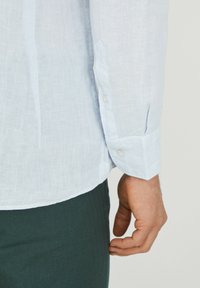Camicia di lino azzurro chiaro con maniche lunghe, polsini con bottoni e tessuto textured. Una mano, appoggiata su pantaloni verde scuro, è parzialmente visibile.