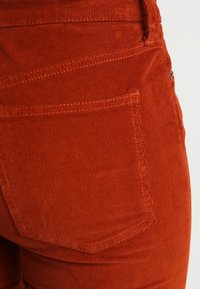Gros plan sur la poche arrière d'un pantalon en velours côtelé orange brûlé avec des coutures visibles et une texture de tissu.