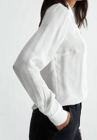 Blusa bianca a maniche lunghe con un motivo sottile, dotata di tasca e polsini con bottoni, abbinata a jeans scuri, vista di lato.