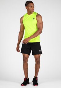 Neon groene mouwloze sportshirt met zwarte shorts. Zwarte sneakers met rode accenten. Glad materiaal, strakke pasvorm, minimaal logo op het shirt.